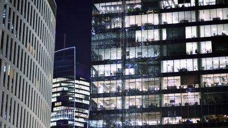 Office building in the evening with lightened workspaces. Modern, glazed skyscraper in financial district. Late hours in the office.の写真素材