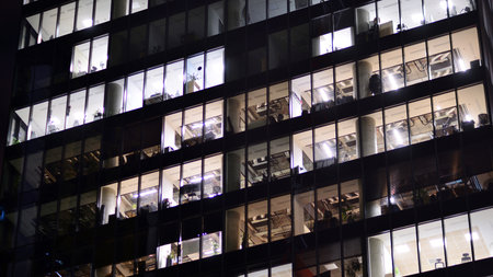 Office building in the evening with lightened workspaces. Modern, glazed skyscraper in financial district. Late hours in the office.の写真素材