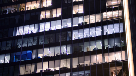 Office building in the evening with lightened workspaces. Modern, glazed skyscraper in financial district. Late hours in the office.の写真素材