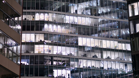 Office building in the evening with lightened workspaces. Modern, glazed skyscraper in financial district. Late hours in the office.の写真素材