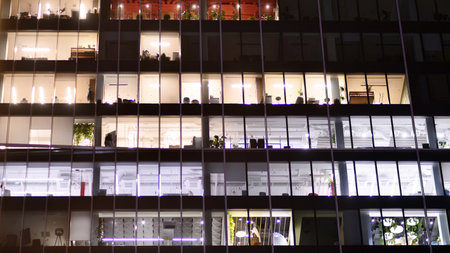 Office building in the evening with lightened workspaces. Modern, glazed skyscraper in financial district. Late hours in the office.の写真素材