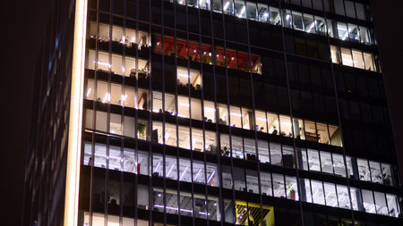 Office building in the evening with lightened workspaces. Modern, glazed skyscraper in financial district. Late hours in the office.の写真素材