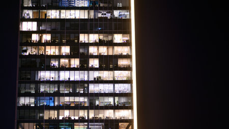 Office building in the evening with lightened workspaces. Modern, glazed skyscraper in financial district. Late hours in the office.の写真素材