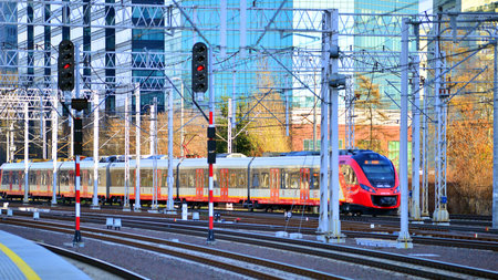 Warsaw, Poland. 28 November 2025. A passenger train on the tracks near the station. Tracks, electrical wires and metal masts against the backdrop of a city skyscrapers on a winterのeditorial素材