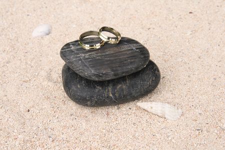 A pair of gold wedding rings placed upon black pebbles on the sand beside a white seashellの写真素材