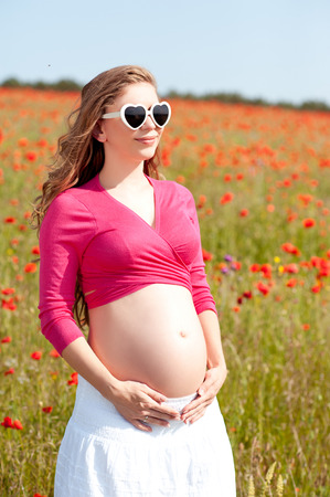 Beautiful pregnant woman resting in poppy field. Wearing sun glasses outdoors. Looking away.の写真素材