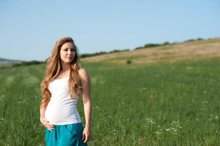 Pregnant woman holding tummy in meadow outdoors. Young adults. Motherhood. Maternity. Looking away.の写真素材