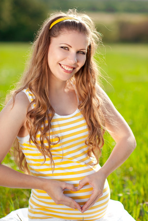 Smiling pregnant woman resting outdoors. Making heart with fingers. Summer time. Motherhood. Maternity. Happiness. 20s.の写真素材