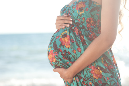 Pregnancy. Pregnant woman holding belly outdoors. Resting at seashore. Wearing floral dress.の写真素材