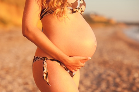 Pregnant woman holding her belly outdoors. Wearing swimsuit at seashore. Summer vacation.の写真素材