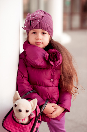 Smiling baby girl 3-4 year old holding toy pet in bag outdoors. Looking at camera. Wearing trendy winter jacket. Childhood.の写真素材