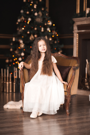 Beautiful kid girl 4-5 year old wearing stylish dress sitting on vintage armchair in room over Christmas tree. Looking at camera. Winter season. Merry Christmas. の写真素材