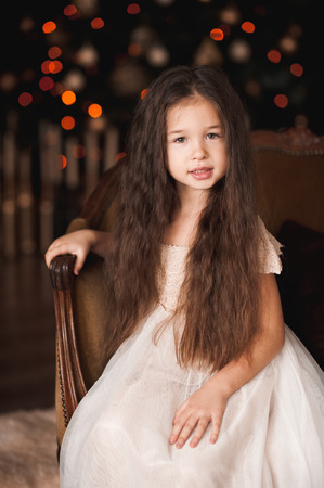 Beautiful baby girl 4-5 year old sitting in armchair over Christmas tree in room. Looking at camera. Holiday season.の写真素材