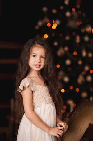 Smiling baby girl 5-6 year old wearing stylish dress standing in room over Christmas tree at night. Looking at camera. Holiday season. の写真素材