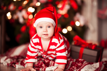 Funny baby 6-8 months old wearing striped santa claus suit and hat crawling on floor over Christmas tree lights. Looking at camera. Merry Christmas. Holiday season. の写真素材