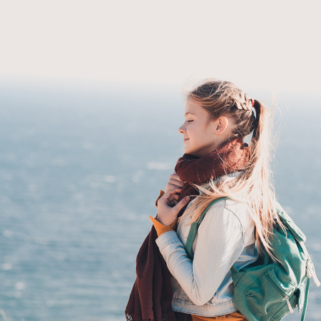 Smiling teen girl 14-16 year old traveling with backpack over sea background. Wearing warm scarf,...