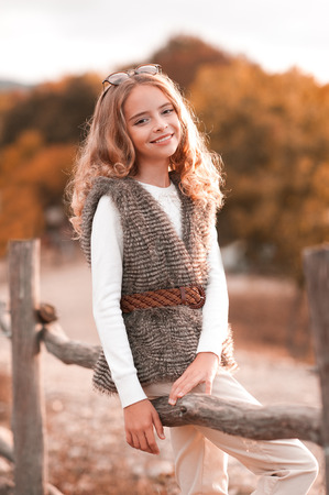 Beautiful blonde teenage girl 13-14 year old wearing fur vest, white knitted sweater and beige pants. Posing at country side. Looking at camera. Autumn season.の写真素材