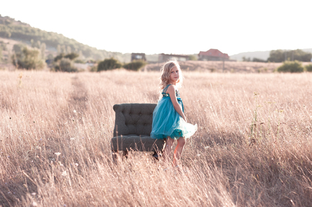 Stylish kid girl 4-5 year old sitting on armchair in meadow wearing trendy dress outdoors. Looking at camera. の写真素材