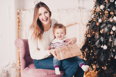 Laughing mother open Christmas presents with her baby girl 1 year old sitting in room with Christmas tree. Looking at camera. Holiday season. Family time.の写真素材