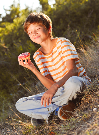 Happy kid boy 12-14 year old eating donut outdoors. Eyes closed. Sunny day.の写真素材