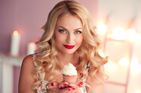 Smiling blonde girl holding creamy cupcake in room closeup. Looking at camera. Bakery. の写真素材
