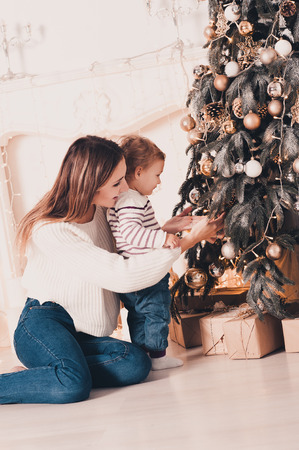 Young mother decorating Christmas tree with baby girl under 1 year old in room. Holiday season. の写真素材