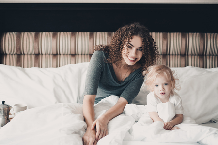 Smiling young mother sitting in bed with baby girl in hotel room. Looking at camera. Good morning. の写真素材