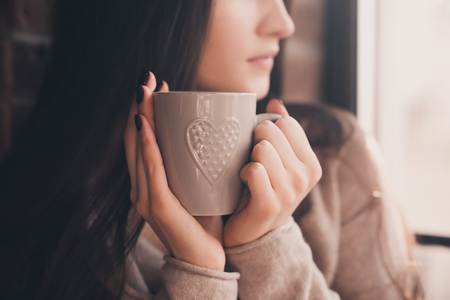 Brunette woman 20-24 year old holding ceramic cup of tea closeup. Looking at window. Good morning. Selective focus. 20s.の写真素材