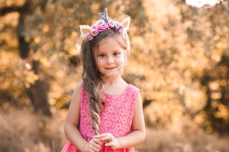 Smiling kid girl 4-5 year old with long hair wearing pink dress and unicorn headband outdoors. Looking at camera. Childhood. の写真素材