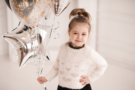 Smiling kid girl 5-6 year old holding silver star balloons wearing knitted sweater with hearts in white room close up. Looking at camera. Birthday party.の写真素材
