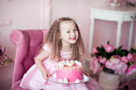 Happy kid girl 4-5 year old sitting in pink armchair with tasty cake in room. Looking at camera. Birthday party. の写真素材