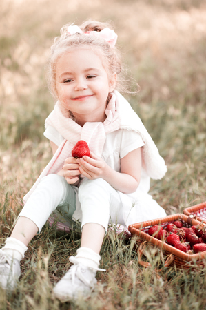 Smiling baby girl 3-4 year old eating strawberries sitting outdoors. Looking at camera. Summer time. の写真素材
