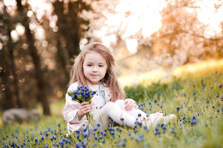 Smiling kid girl 4-5 year old holding blue flowers outdoors. Sitting in park wearing spring clothes. Looking at camera. Childhood. の写真素材