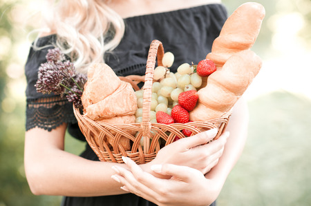 Girl holding basket with food: fresh bread, strawberry, grapes, croissant in park. Prepare for picnic. の写真素材