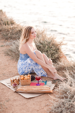 Blonde beautiful girl 22-24 year old having picnic with fresh bread, fruits, cakes and lemonade outdoors. Looking awat ar sea. Good morning. Summer season. 20s.の写真素材