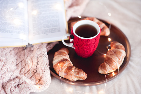 Knitted cup of black tea with homemade croissants and open book staying on wooden tray in bed closeup. Winter season. Good morning.の写真素材