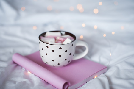 Cup of coffee with marshmallow on open notebook in bed over Christmas lights. Good morning. Tasty breakfast.の写真素材