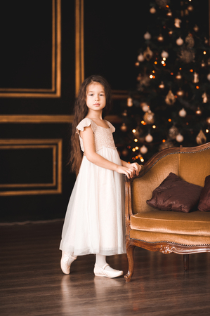 Cute baby girl 4-5 year old wearing stylish white dress posing over Christmas lights in room. Looking at camera. Holiday season.の写真素材