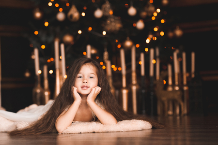 Funny baby girl 5-6 year old lying under Christmas tree  in room. Looking at camera. Winter season.の写真素材