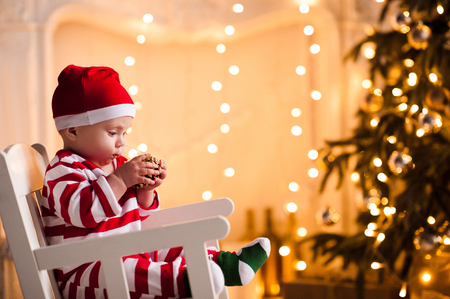 Baby 1 year old wearing santa claus suit sitting in rocking chair over Christmas tree and lights on background in room. Holiday season.の写真素材