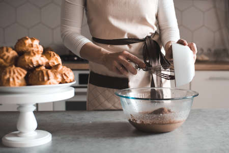 Young woman 25-30 year old cooking homemade chocolate cake at home standing infront of kitchen table mixing batter in bowl closeup. Looking at camera. 20s.の写真素材