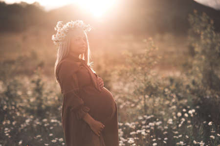 Pregnant woman wearing stylish dress and chamomile wreath resting in meadow outdoors over flower background and sunset light. Selective focus on tummy. Healthy lifestyle. Motherhood. Summer season.の写真素材