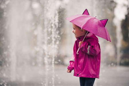 Laughing kid girl 4-5 year old holding pink umbrella wearing raincoat over rain at background closeup in park. Childhood. Autumn season.の写真素材
