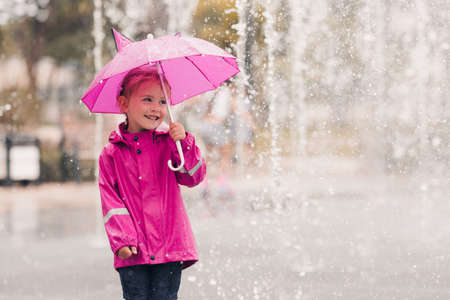 Laughing kid girl 4-5 year old holding pink umbrella wearing raincoat over rain at background closeup in park. Childhood. Autumn season.の写真素材