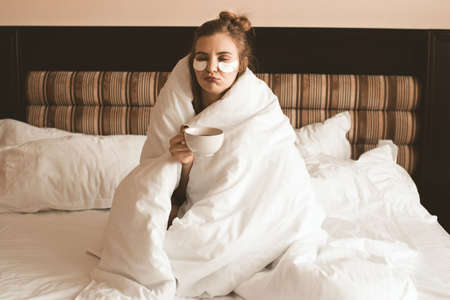 Girl wrapped in blanket with eye patches drinking coffee sitting in cozy bed try to wake up closeup. Good morning. Quarantine Social isolation.の写真素材