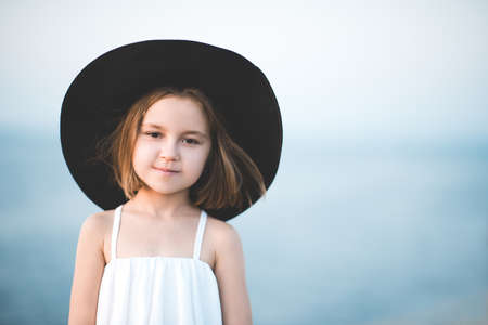 Smiling child girl 4-5 year old wearing big black hat and white top posing over blue sea background close up. Looking at camera. Summer travel season.の写真素材