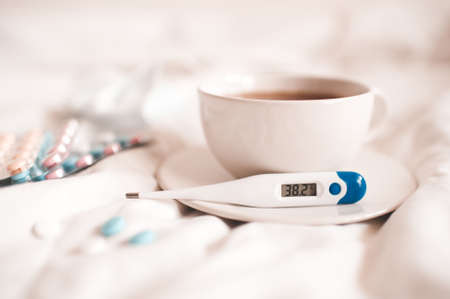 Thermometer with high temperature on plate and cup of tea with pills closeup in bed. Good morning. Illness concept. Social isolation.の写真素材