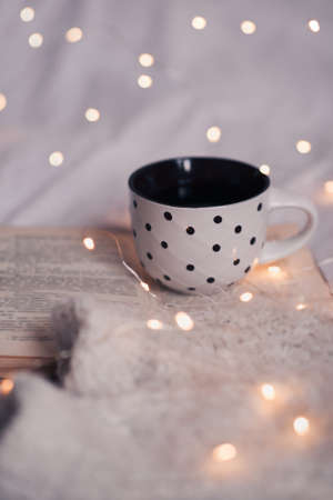 Cup of coofee staying on open book with knitted sweater over glowing lights closeup. Good morning. Breakfast time. Winter season.の写真素材