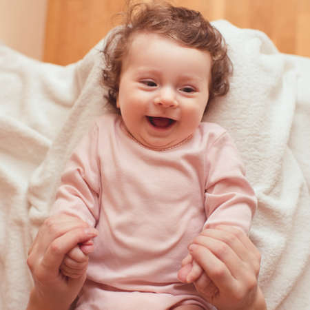 Happy baby with curly hair lying in bed holding mother hands cloeup. Top view. Motherhood. Maternity. Childhood.の写真素材