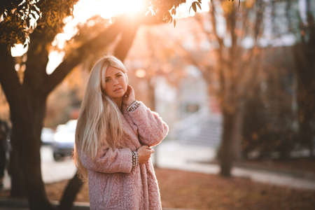 Stylish woman 20-24 year old with long hair wearing pink winter coat leaning on tree in park close up. Lokking at camera. 20s.の写真素材